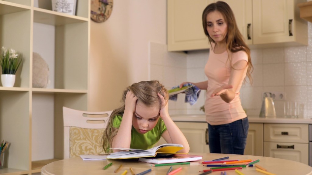 Eine junge Mutter schimpft mit ihrer jungen Tochter, die an ihren Hausaufgaben sitzt.