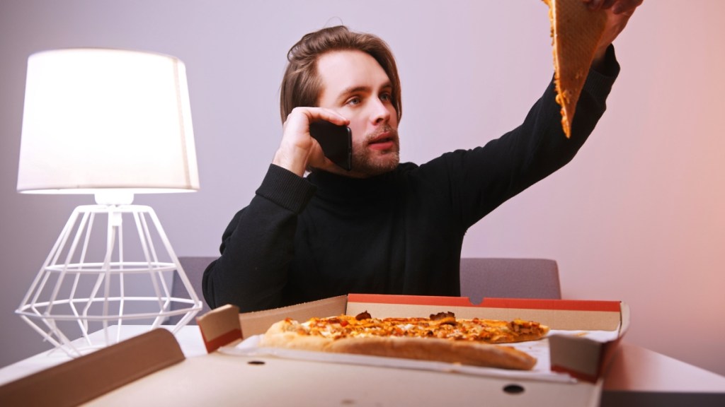 Ein junger Mann mit schwarzem Pullover hält beim Telefonieren ein Stück Pizza in der Hand.