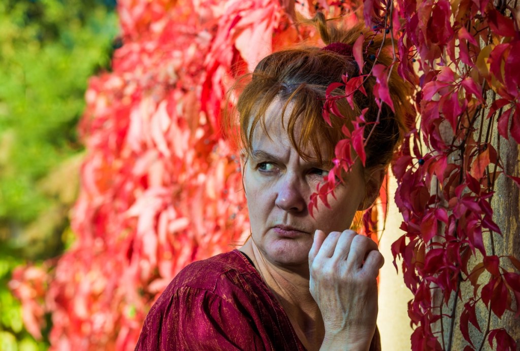 Eine Frau steht vor einer Hecke und schaut argwöhnisch über ihre Schulter.
