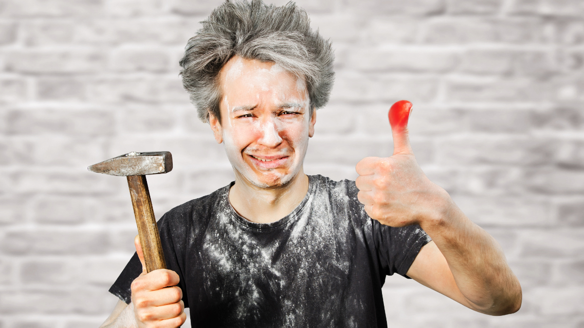 Ein schmutziger Handwerker mit einem Hammer in der Hand hält einen geschwollen Daumen hoch und macht ein schmerzverzerrtes Gesicht.