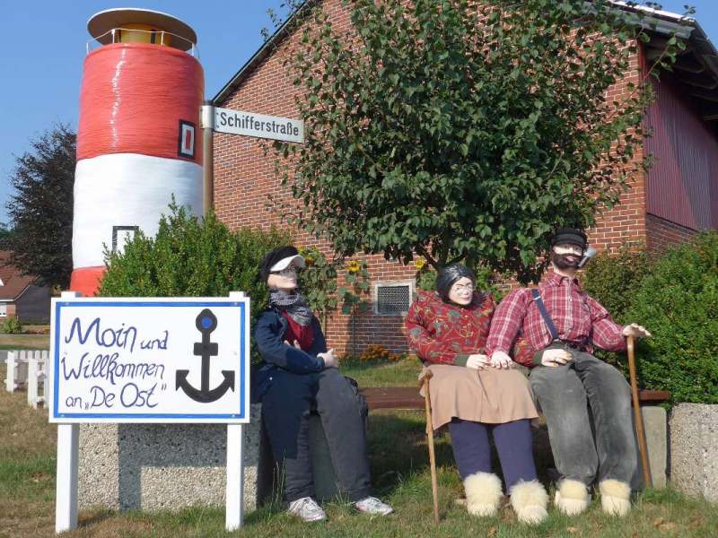 Drei Puppen sitzen auf einer Bank vor einem Leuchtturm in Ostfriesland.