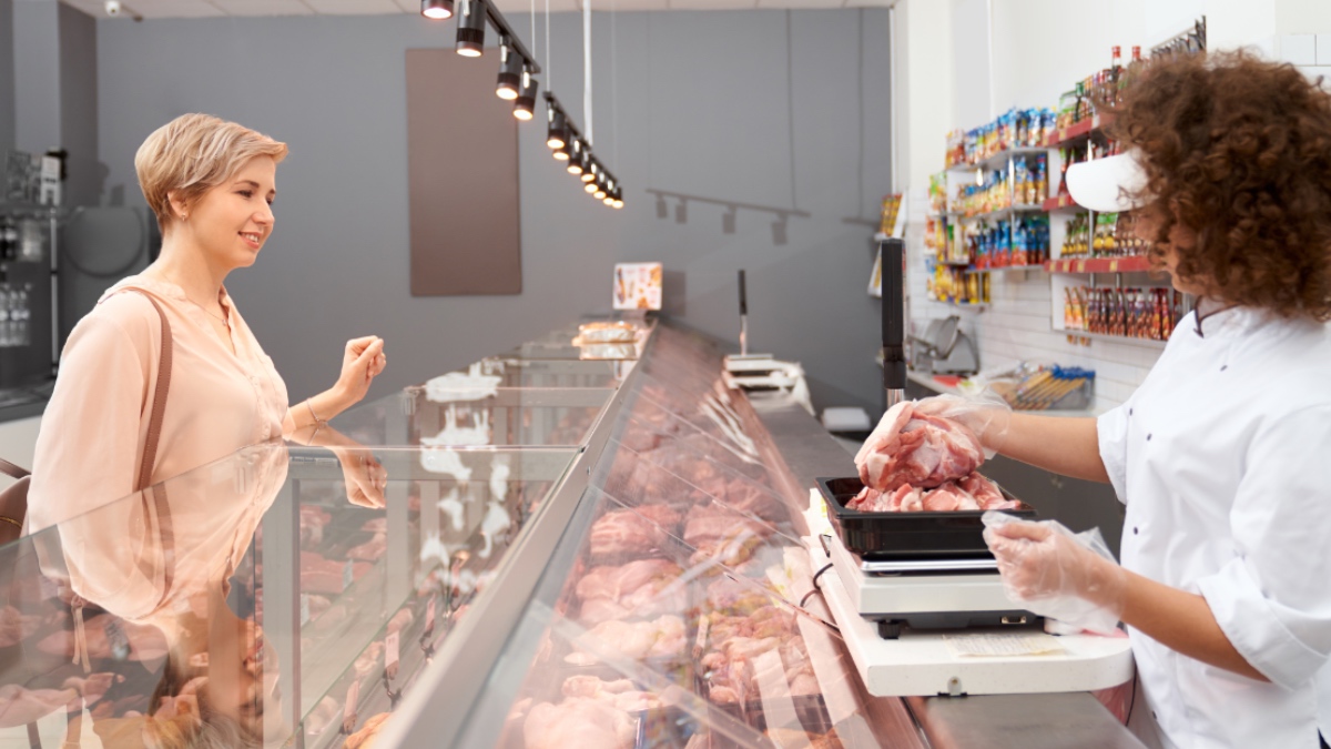 Seitenansicht einer jungen lächelnden Frau mit lockigem Haar, die einer positiven Dame rohes Schweinefleisch vorführt.